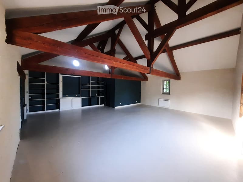 Empty room with exposed wooden beams on the ceiling, white walls, grey floor, built-in shelving unit, and a small window.