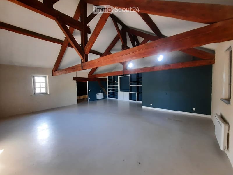 Empty room with exposed wooden beams, white walls, and a single window. The floor is smooth and grey, and there is a small built-in shelving unit in the corner.