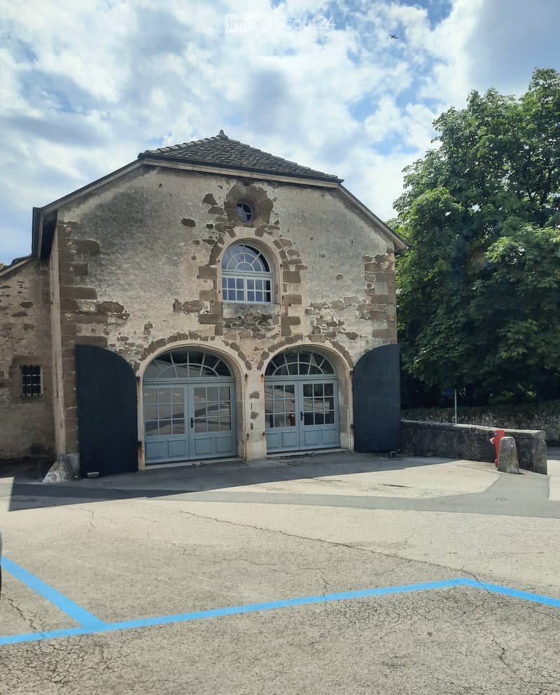 Stone building with arched doors and windows, surrounded by greenery and a paved area.