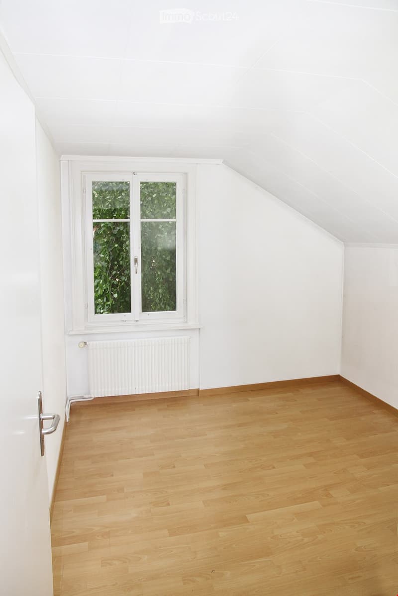Empty room with wooden flooring, white walls, and a large window with a view of greenery. The room features a radiator below the window and a door on the left side.