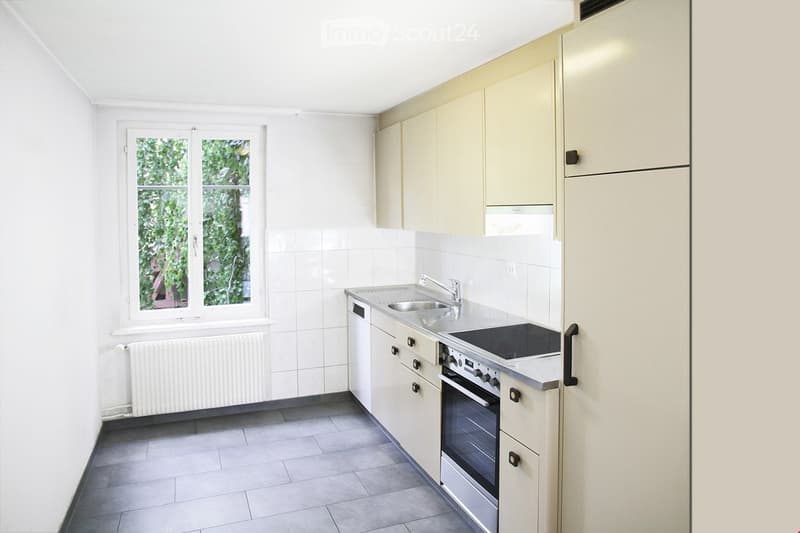 Modern kitchen with white cabinets, stainless steel sink, oven, and refrigerator. Features a window with a view of greenery and a radiator below.