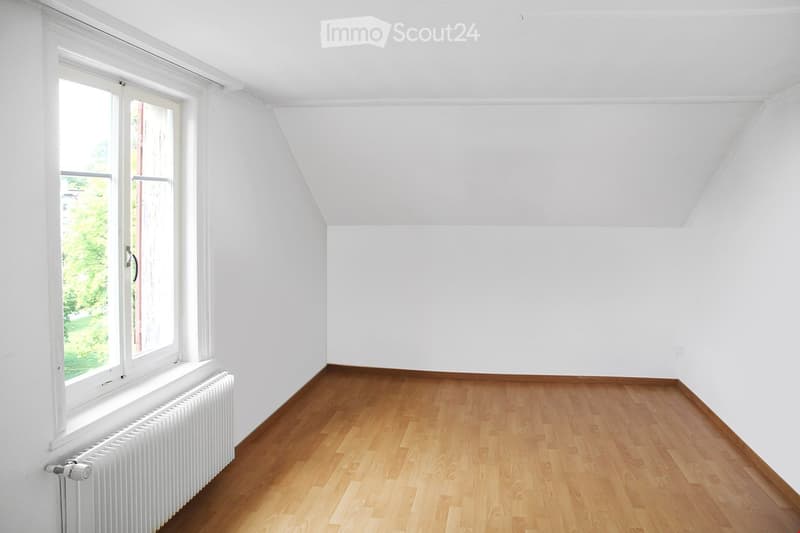 Completely empty room with white walls, wooden floor, and a large window with a radiator below it.