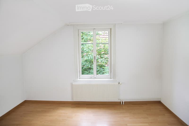 Empty room with wooden flooring, white walls, a single window with white frame, and a white radiator below the window.