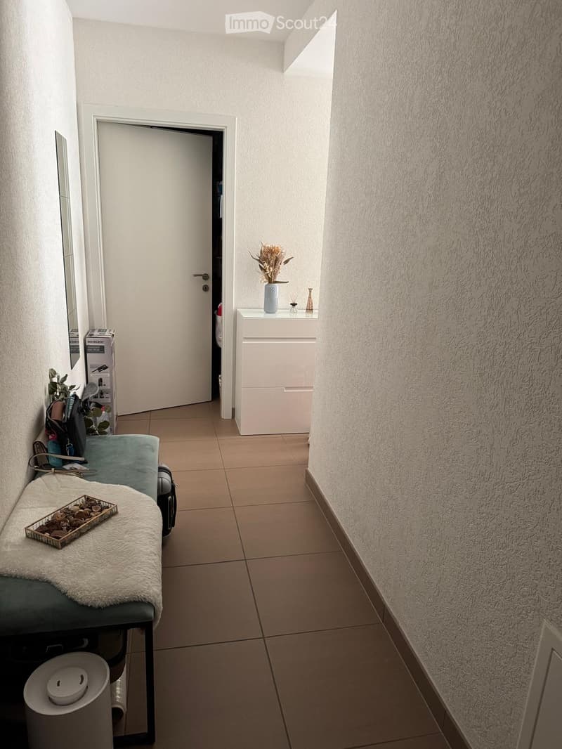 A narrow hallway with tiled flooring, white walls, and a door leading to another room. There is a small white cabinet with a vase and decorative items on top, and a bench with a white blanket and a small tray on it.