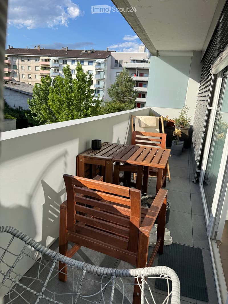 Balcony with wooden table, chairs, bench, and small potted plants, overlooking a cityscape with buildings and trees.