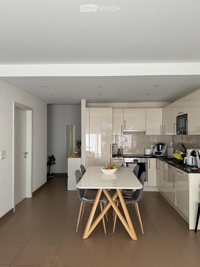 Modern kitchen with light-colored cabinets, a white dining table with six chairs, and various appliances including a refrigerator, microwave, oven, coffee maker, and toaster. A bowl of fruit is placed on the table.