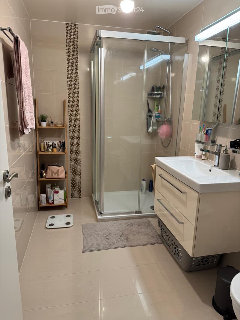 Modern bathroom with beige tiles, a glass-enclosed shower, a sink with a mirror, a towel on a rack, a small shelf with toiletries, and a bathroom scale on the floor.