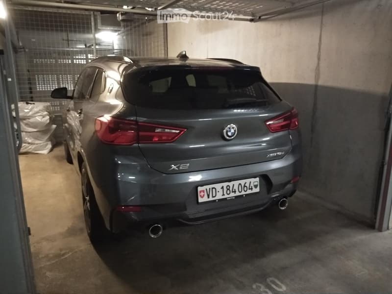 Underground parking space with a grey BMW X2 SUV parked inside, concrete walls and ceiling, and a metal gate visible on the left side.
