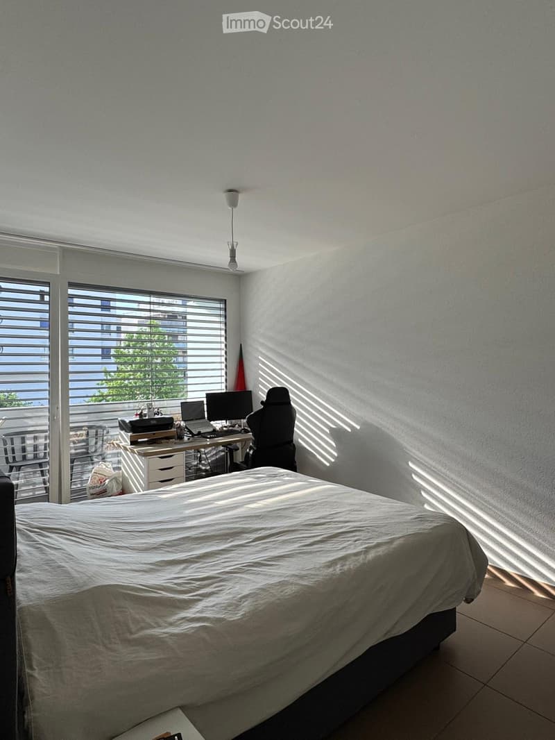 A well-lit bedroom with a bed, desk, and chair, featuring large windows with blinds and a view of the balcony.