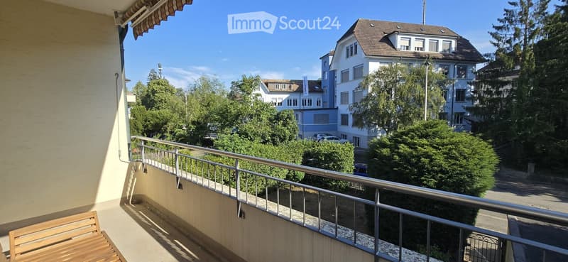 Balcony with wooden bench, metal railing, overlooking a street with trees and buildings.