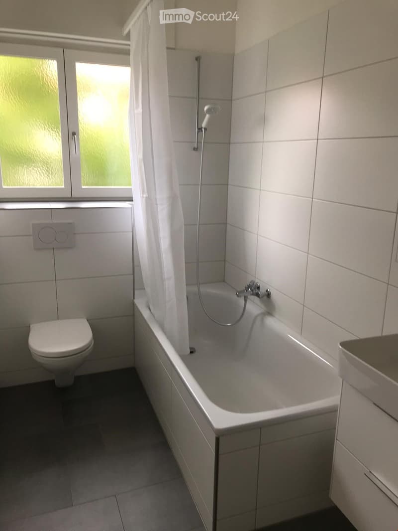 Modern bathroom with a bathtub, toilet, and showerhead, featuring white tiles and a frosted window.