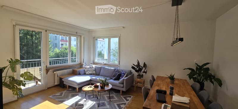 Bright living room with large windows and balcony access, featuring a grey sectional sofa, coffee table, side table, dining table, chairs, and several potted plants.