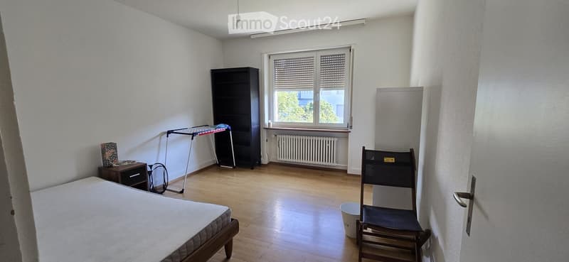 A furnished bedroom with a bed, a wooden chair, a bookcase, a small table, a radiator, and a stool. The room has wooden flooring and white walls.