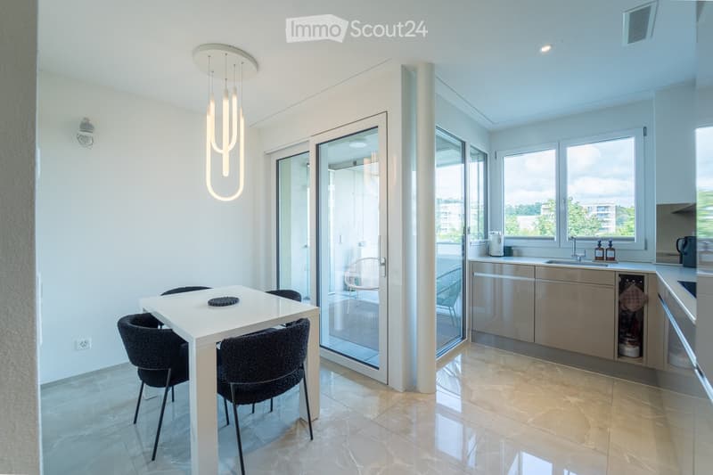 Modern, open-plan kitchen and dining area with white walls, light-colored tiles, and large windows. Features a white dining table with four black chairs, a sleek kitchen counter with cabinets, a sink, and a dishwasher. A pendant light hangs above the dini