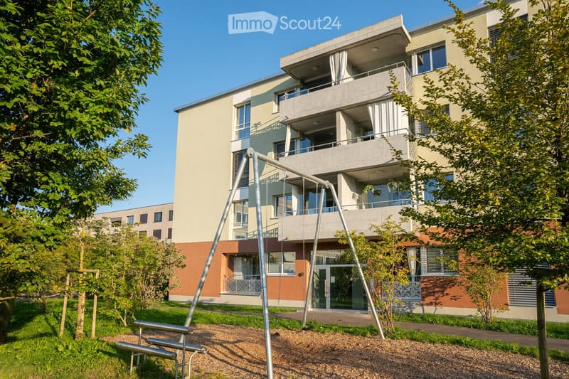 Modern multi-story residential building with balconies, surrounded by greenery and playground equipment.