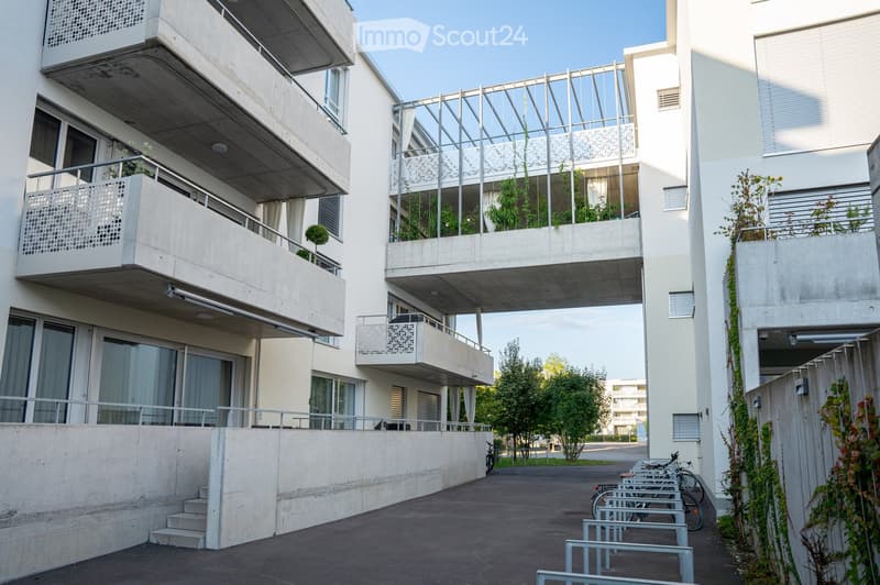 Modern multi-story residential building with concrete balconies, large windows, and greenery. Bicycles are parked in the foreground.