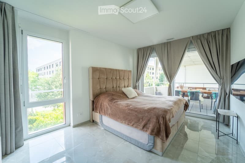 Modern bedroom with a large bed, beige headboard, and a balcony with outdoor furniture visible through large windows.