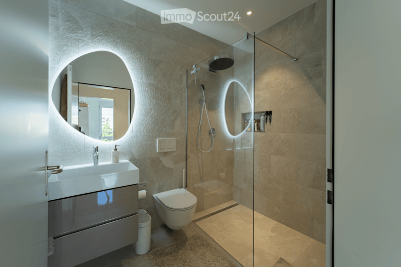 Modern bathroom with a white sink, oval mirror, toilet, glass-enclosed shower, and built-in storage. Features include a sleek faucet, modern lighting, and a small trash bin.
