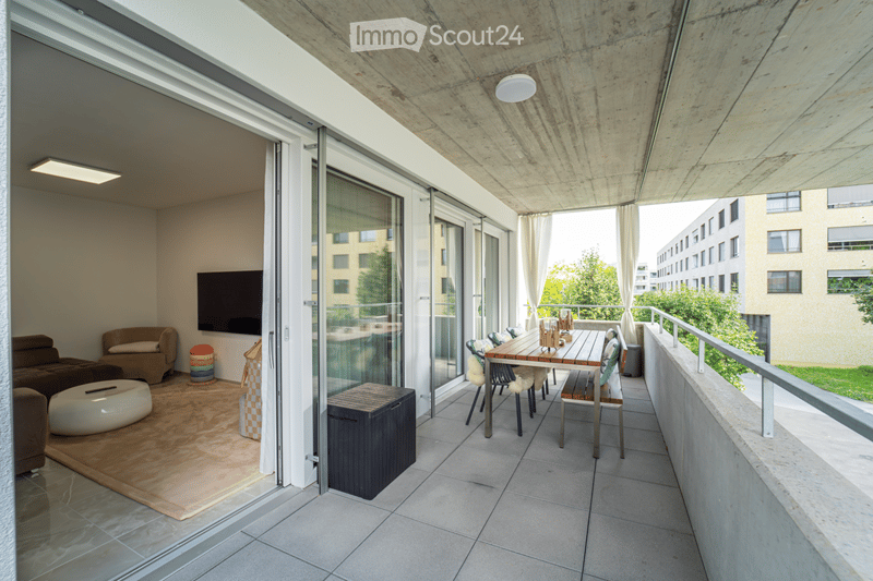 Modern living room with a large balcony, featuring a sofa, ottoman, round stool, and a dining table with chairs on the balcony.