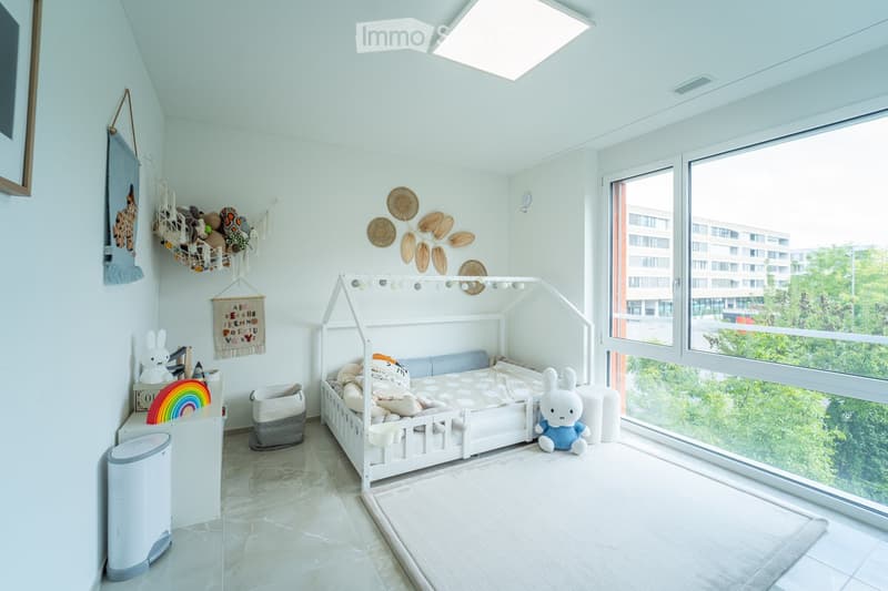 Bright, modern children's bedroom with a white wooden bed, colorful pillows, and a view of greenery outside. Features include a hanging toy basket, a small table with a rainbow decoration, and a white storage basket.
