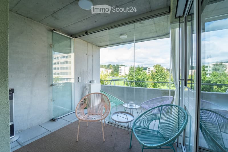 Modern balcony with large glass windows, offering a view of greenery and buildings. Furnished with colorful wicker chairs and small tables.