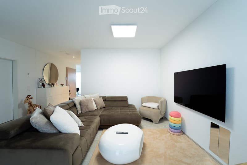 Modern living room with a brown sectional sofa, white stool, beige armchair, large flat-screen TV, and decorative items including a mirror and a stack of colorful cushions.