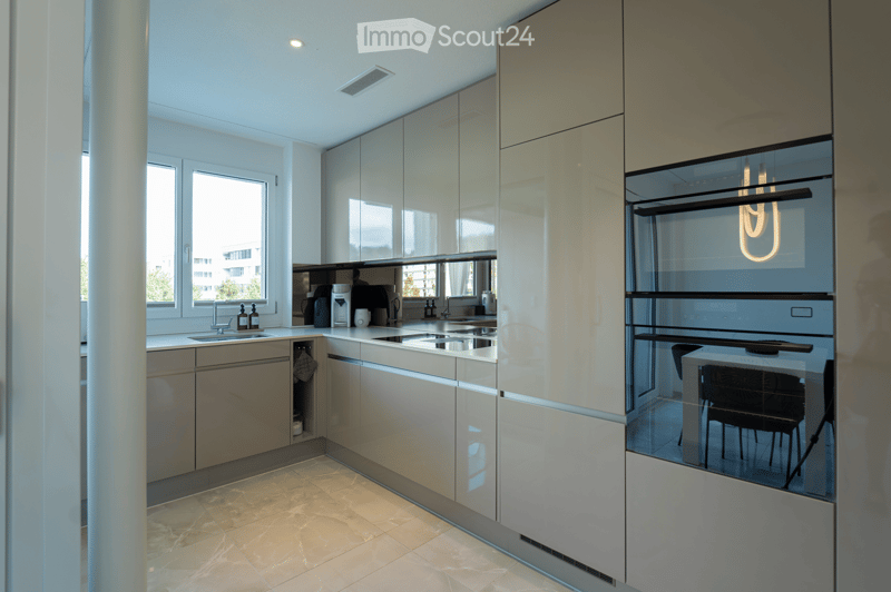 Modern kitchen with sleek, light-colored cabinets, stainless steel appliances, and a large window providing natural light. Features a sink, coffee maker, and an oven with a glass door.