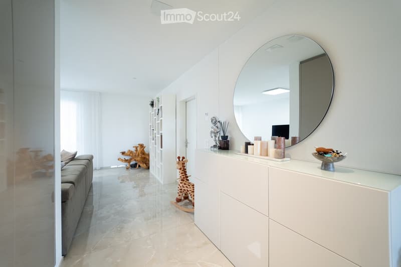 Modern living room with a grey sofa, a large round mirror, a white table with candles, a rocking horse, and a decorative sculpture.