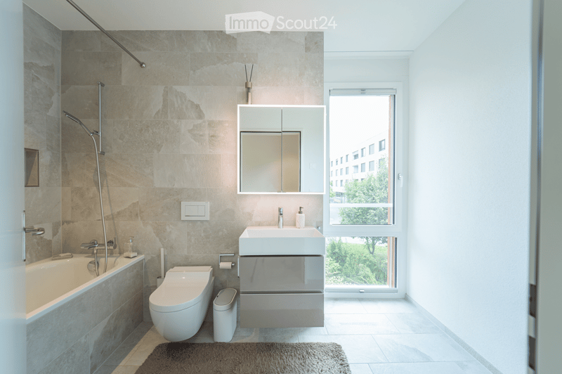 Modern bathroom with beige marble tiles, a bathtub, toilet, sink, mirror, and a large window offering a view of greenery.