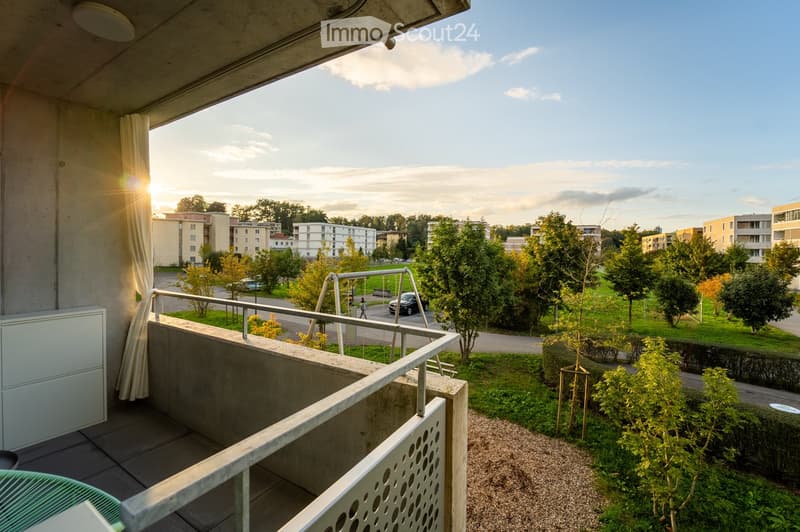 Balcony with a view of a residential area, trees, and a playground. The balcony has a metal railing and a tiled floor.