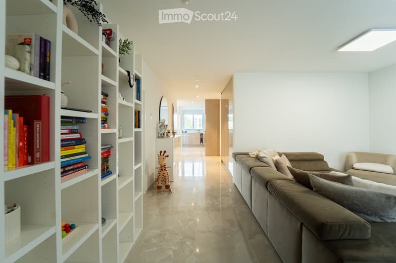 Modern living room with a beige sofa, matching chair, white bookshelf filled with books, a small giraffe toy, and a mirror on the wall.