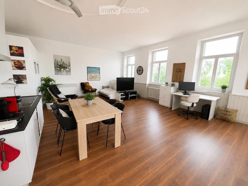 A modern, open-plan living and working space with wooden flooring, large windows, a dining table with chairs, a sofa, a bed, a TV, a desk with a computer, and various decorative items including plants and artwork.