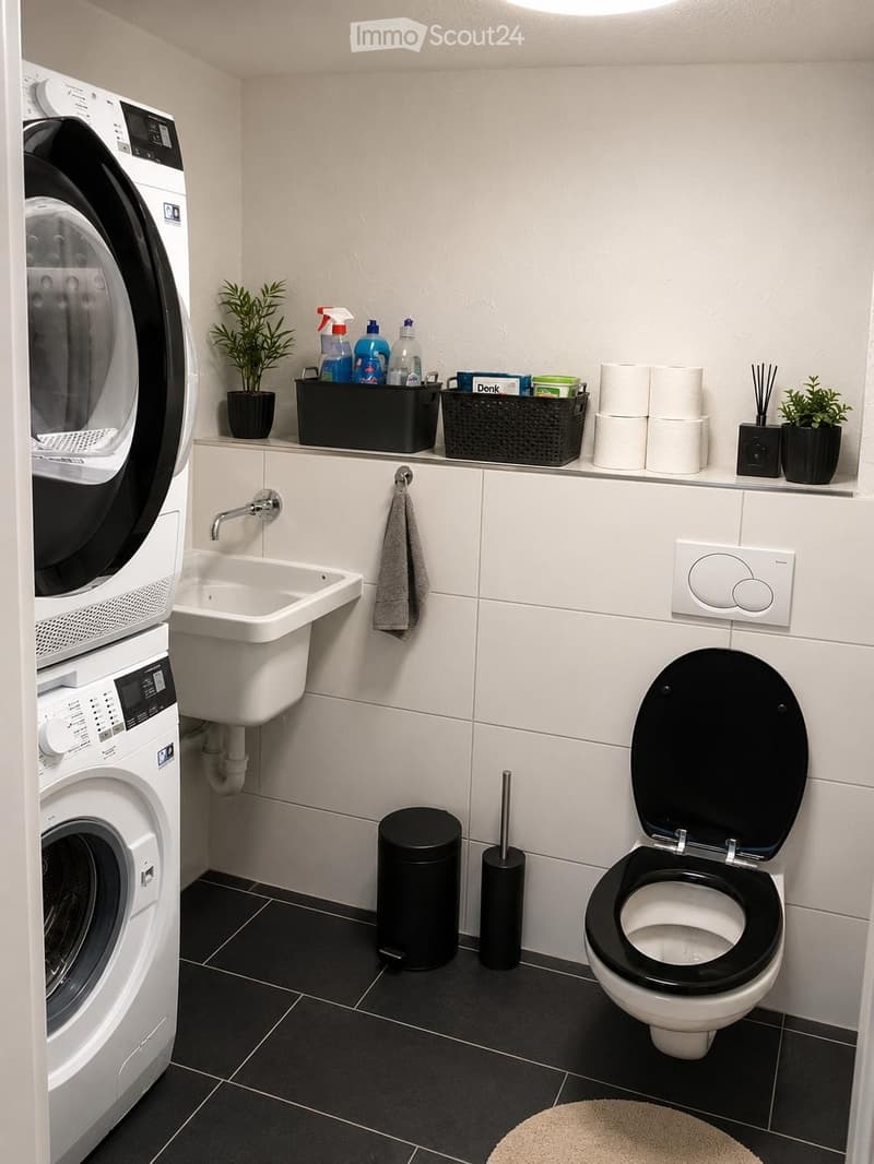 Modern bathroom with white tiles, black floor tiles, a toilet, sink, washing machine, and dryer. Includes a trash can, toilet brush, towel, small plant, cleaning supplies, and toilet paper on a shelf.
