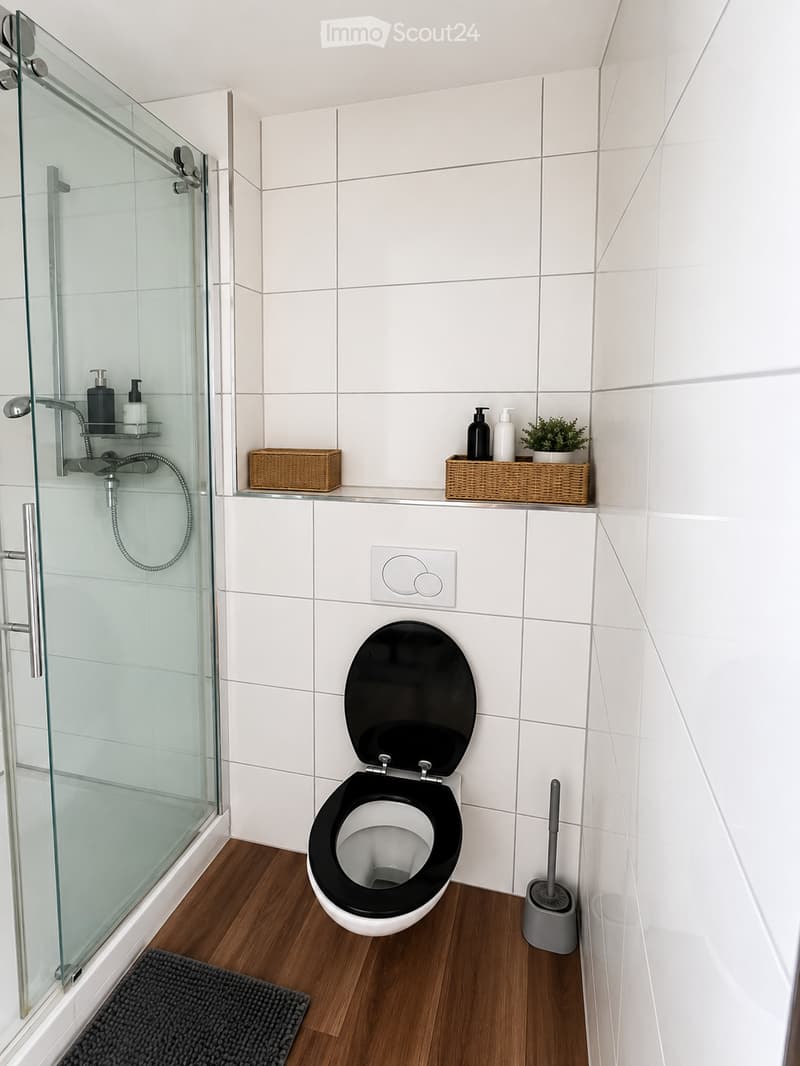 Modern bathroom with white tiled walls, a black toilet seat, a glass-enclosed shower, and a small wicker basket on the shelf holding toiletries and a small plant.