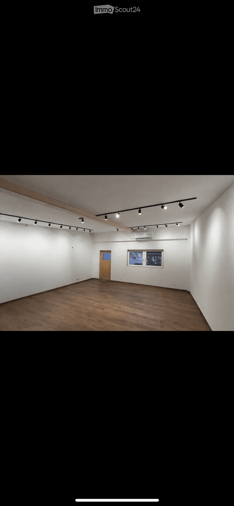 Empty room with wooden flooring, white walls, a single door, and a window with a view of a vehicle outside. Track lighting is mounted on the ceiling.