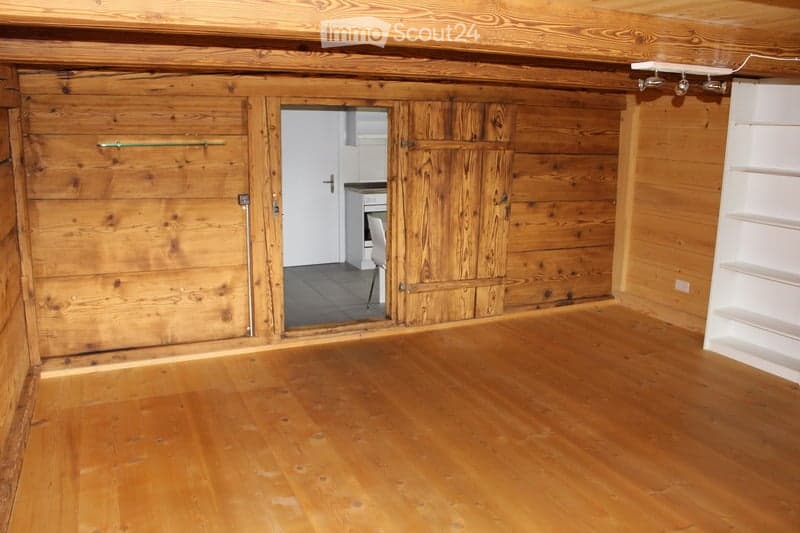 Empty room with wooden walls and floor, featuring a large wooden door and a small adjacent room visible through the doorway.
