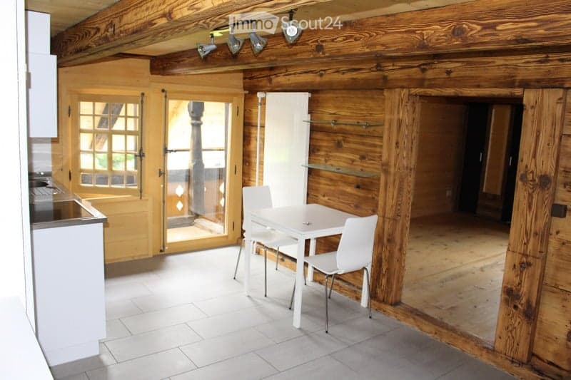 A rustic wooden interior with a white dining table and chairs, a kitchen counter, and large wooden doors leading outside.