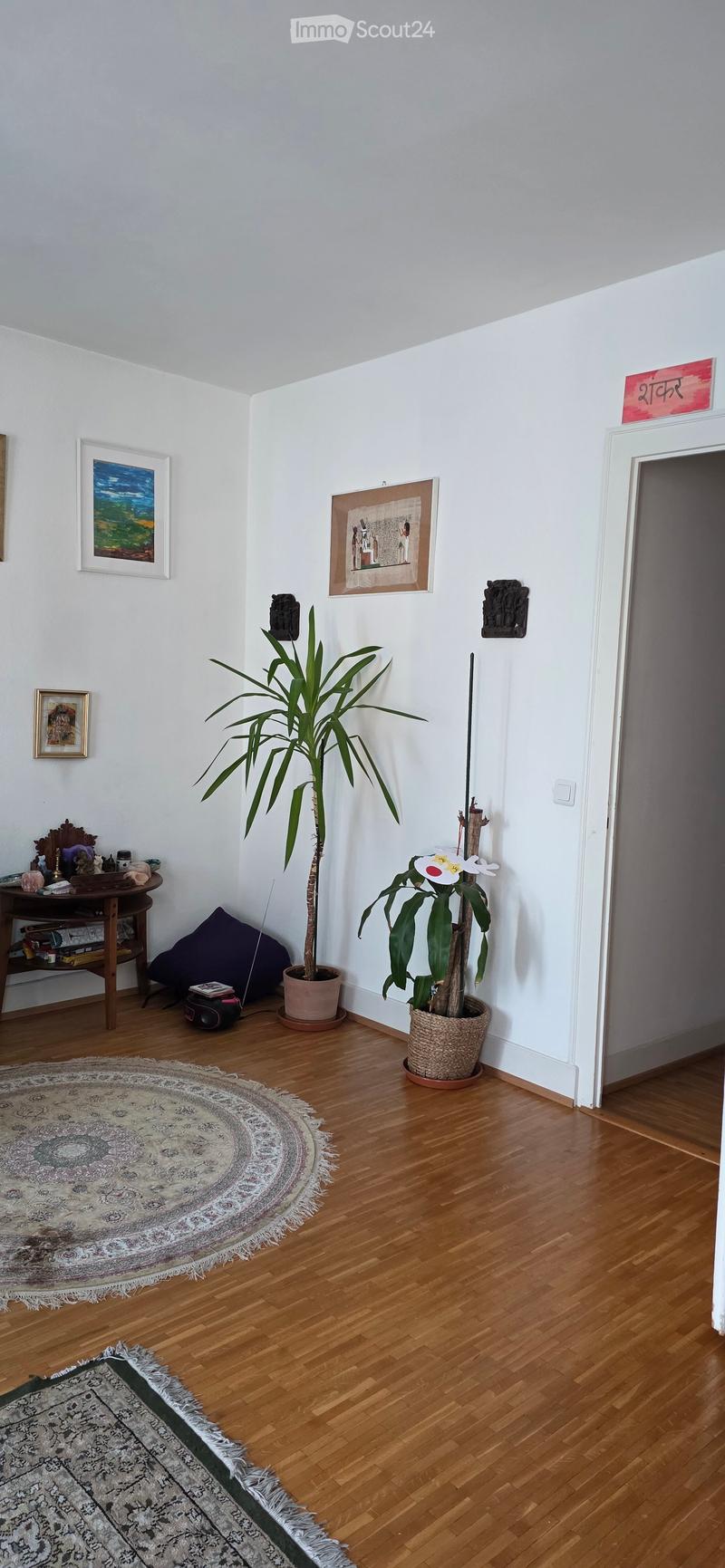 A small, well-lit room with wooden flooring, two potted plants, a round wooden table with a chair, and several decorative items including picture frames and a small shelf with books and objects.