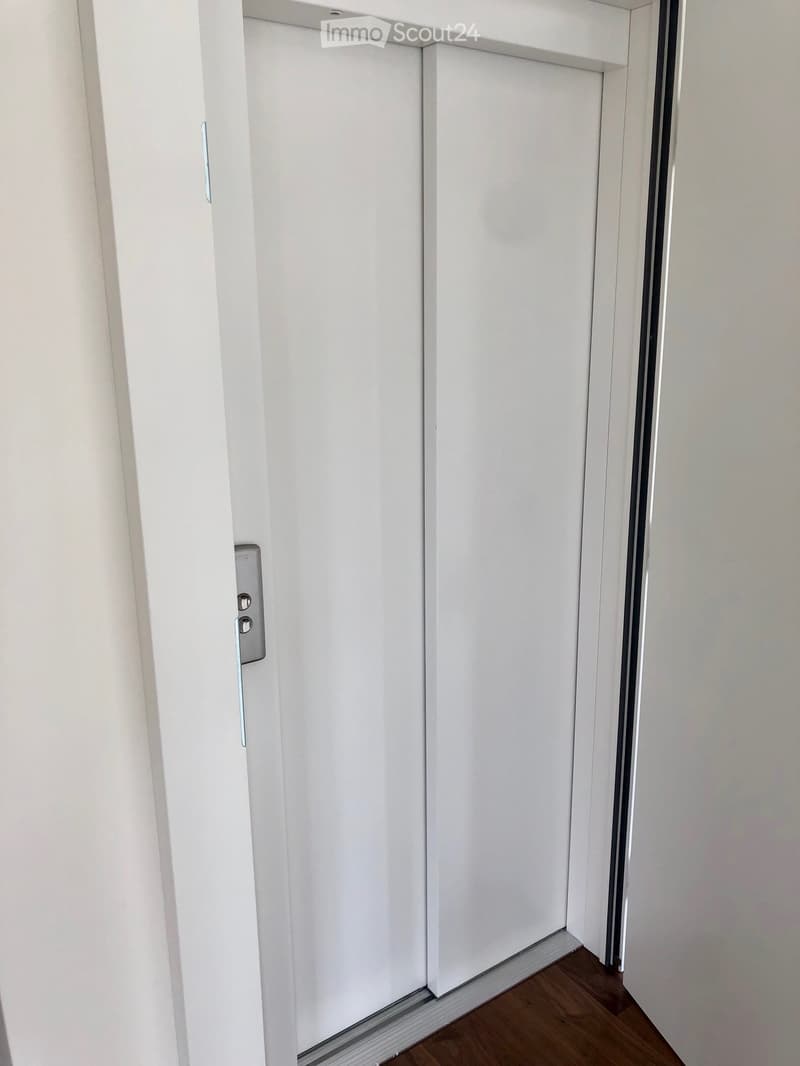 A narrow hallway with white walls, wooden flooring, and a closed white door with a silver handle and lock.