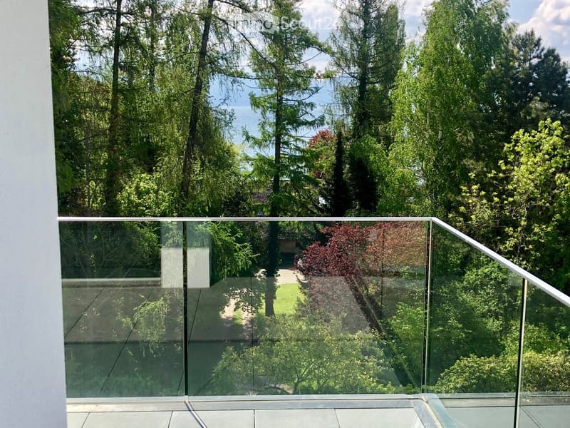 Modern glass balcony with a sleek metal railing, overlooking a lush green garden and trees.