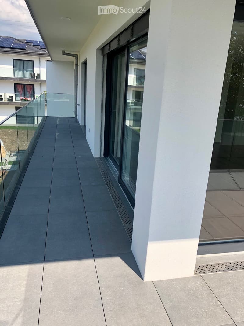 Modern balcony with grey tiled floor, glass railing, and large windows overlooking the building and surrounding area.