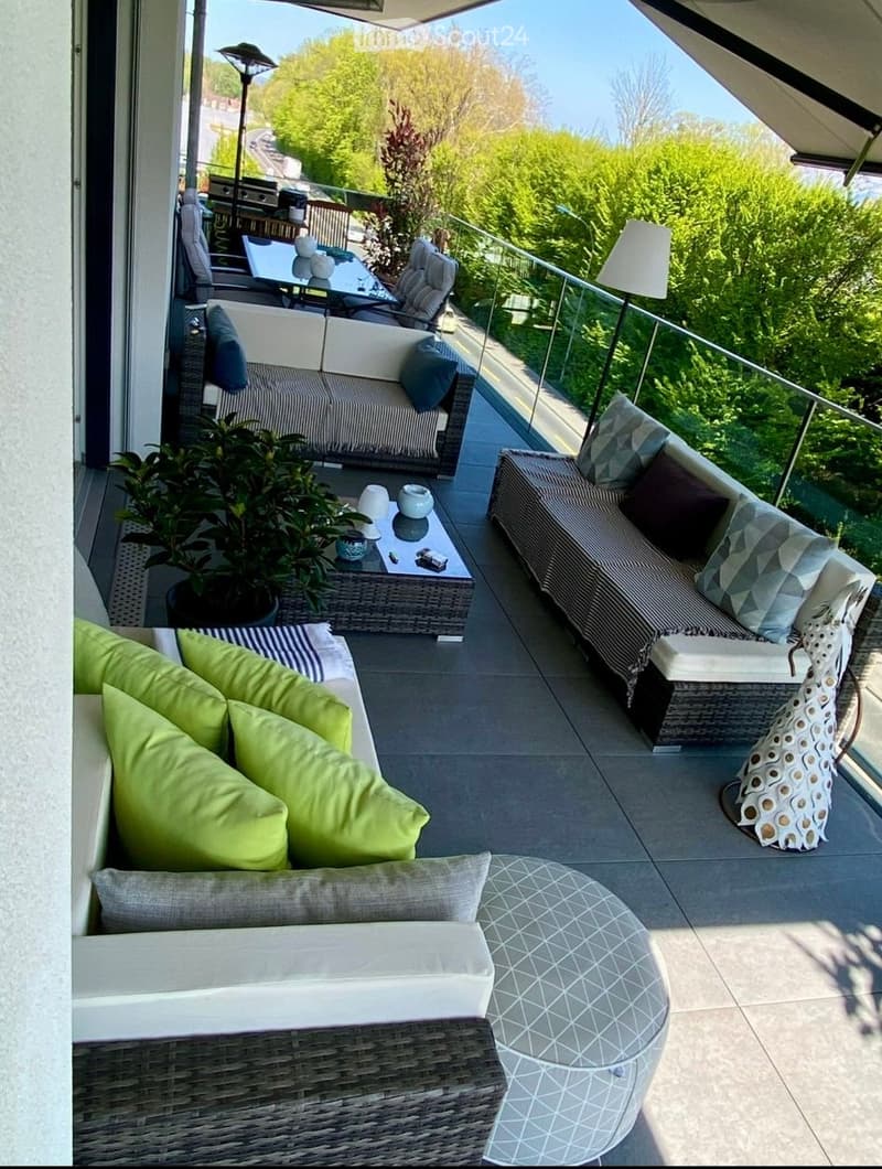 Balcony with modern outdoor furniture including sofas, a table, chairs, and a potted plant, overlooking a green view.