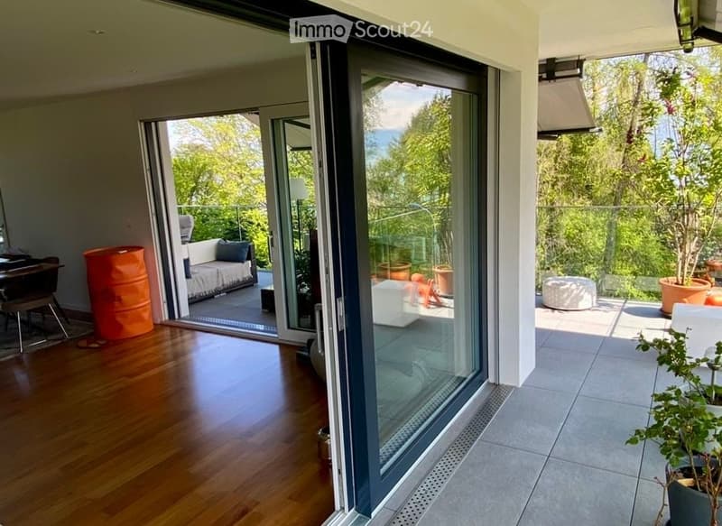 Modern living room with large sliding glass doors opening to a balcony, wooden flooring, white sofa, several stools, and potted plants on the balcony.
