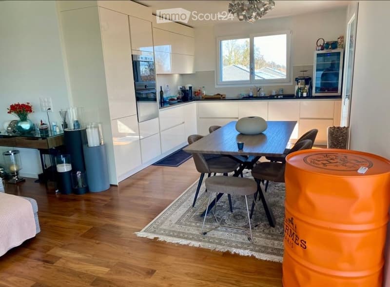 Modern kitchen and dining area with light-colored cabinetry, a central dining table with chairs, and a sleek oven. The space features wooden flooring, a patterned rug, and decorative items including a vase and candles.