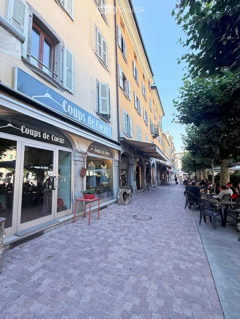 A charming pedestrian street lined with colorful buildings, featuring a clothing store named "Coups de Coeur" on the left and outdoor seating for a café on the right. The street is paved with cobblestones and has trees lining the far side.