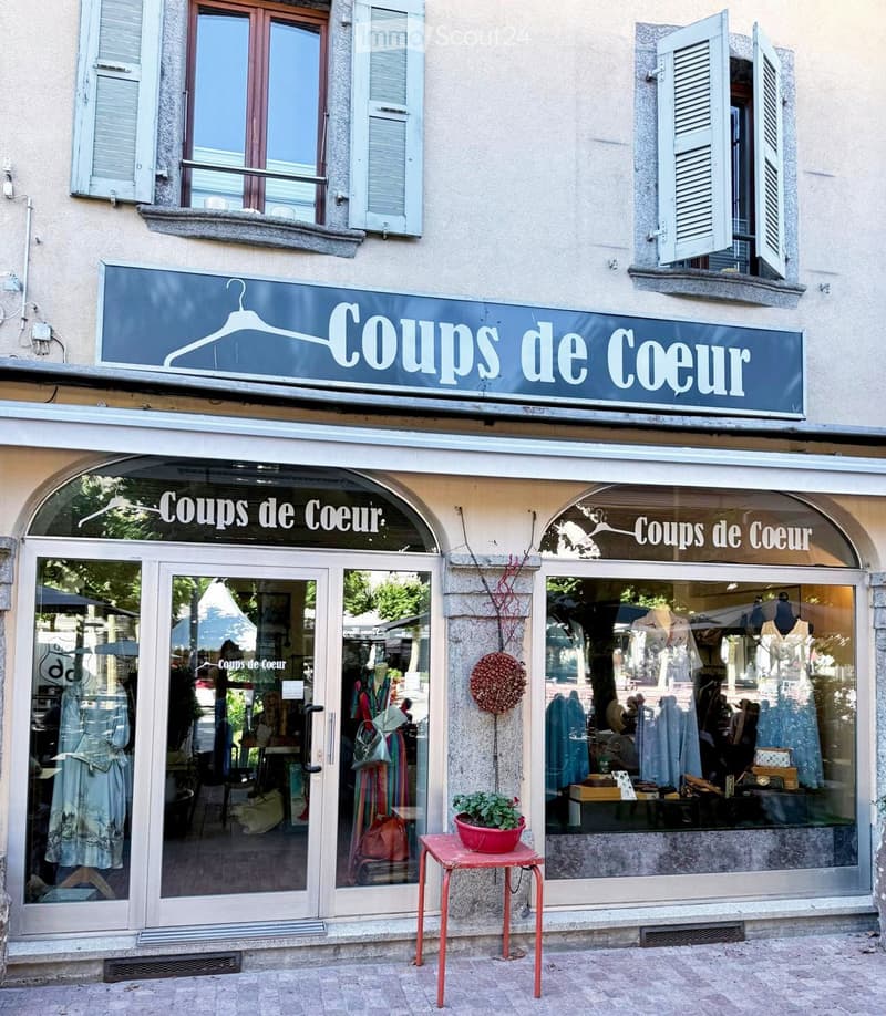 Facade of a clothing store named "Coups de Coeur" with large glass windows displaying mannequins and clothing, a small red table with a potted plant, and a hanging decorative ball.
