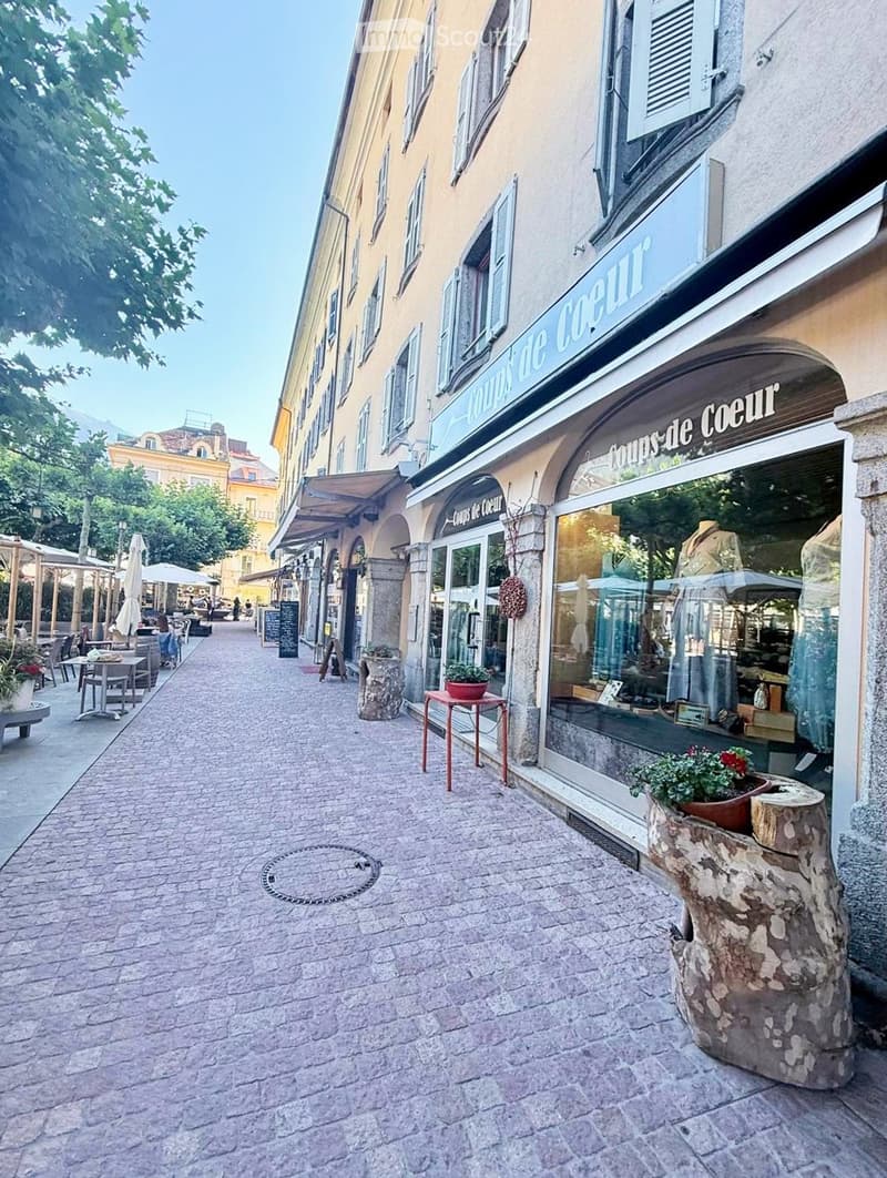 A charming pedestrian street lined with historic buildings, featuring a shop named "Coups de Coeur" with a display window, outdoor seating, and potted plants.
