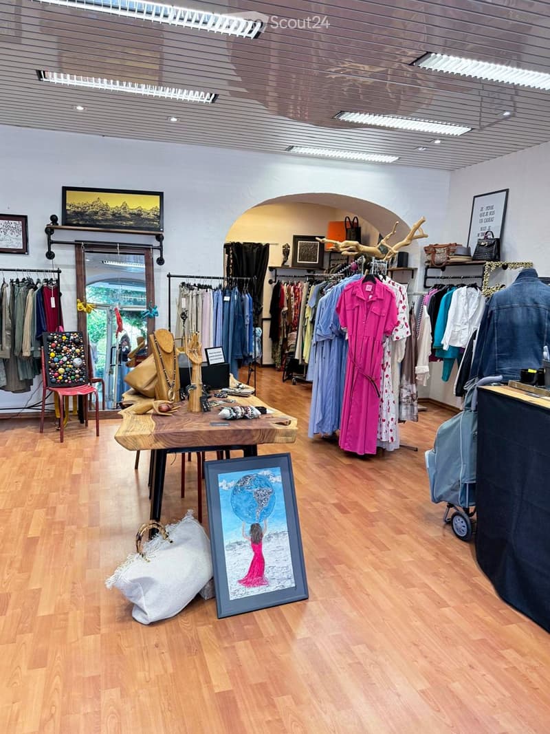 A well-lit clothing boutique with wooden flooring, clothing racks, a wooden table with accessories, and various decorative items including picture frames and a mirror.