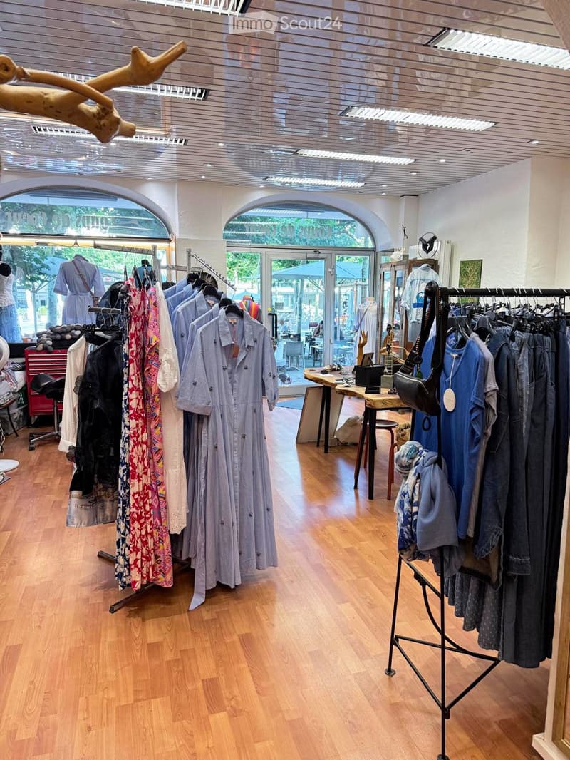 A well-lit clothing store with wooden flooring, multiple clothing racks displaying dresses, shirts, and other garments, a small table with accessories, and a stool.