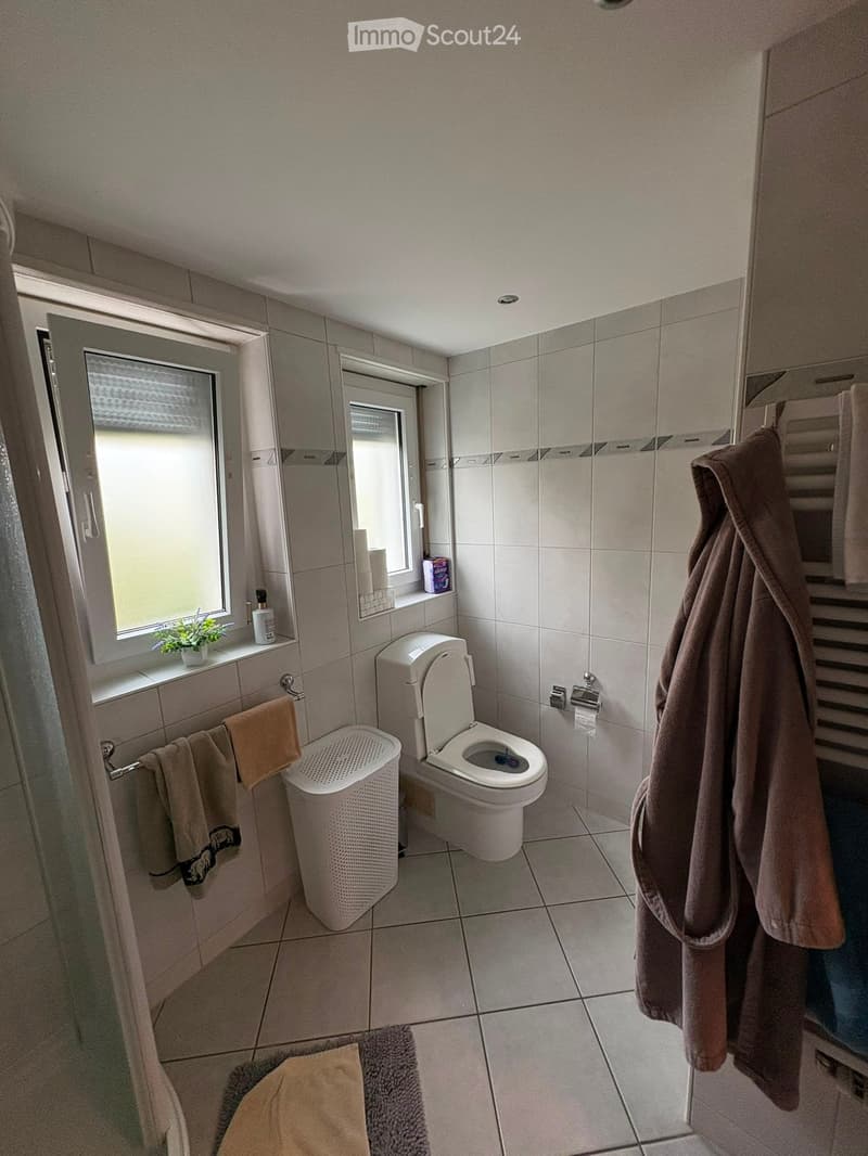 Bathroom with a toilet, towel rack, towels, trash bin, bathrobe, small plant, and bottle on the windowsill.